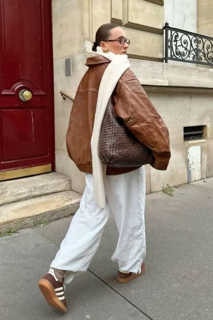 Fashionable woman in a brown leather jacket and white pants walks on a city street in stylish casual attire.