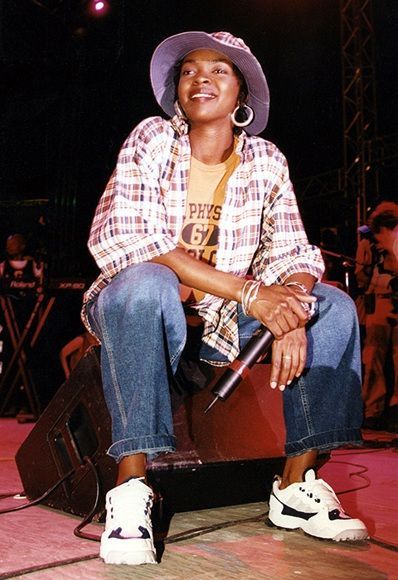 Young woman on stage, wearing a plaid shirt and jeans, holding a microphone, performing at a live music event.