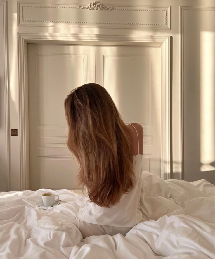 Woman with long hair in cozy white bed, sipping coffee, enjoying morning sunlight. Elegantly styled room interior.