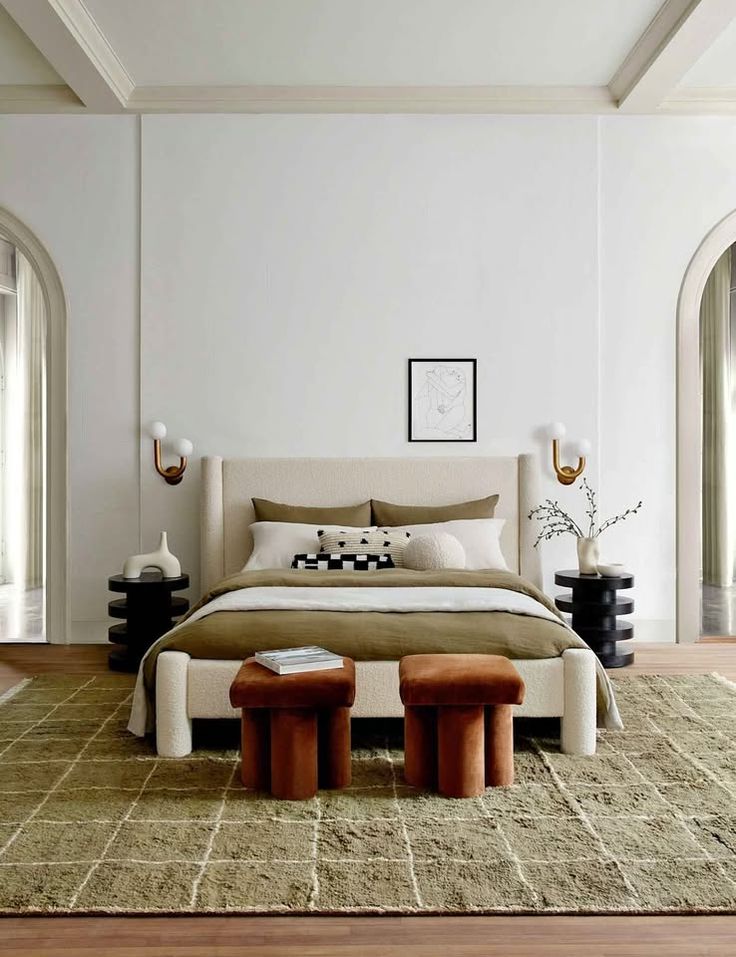 Modern bedroom with beige tones, cozy bed, two brown stools, and stylish decor elements on a patterned rug.
