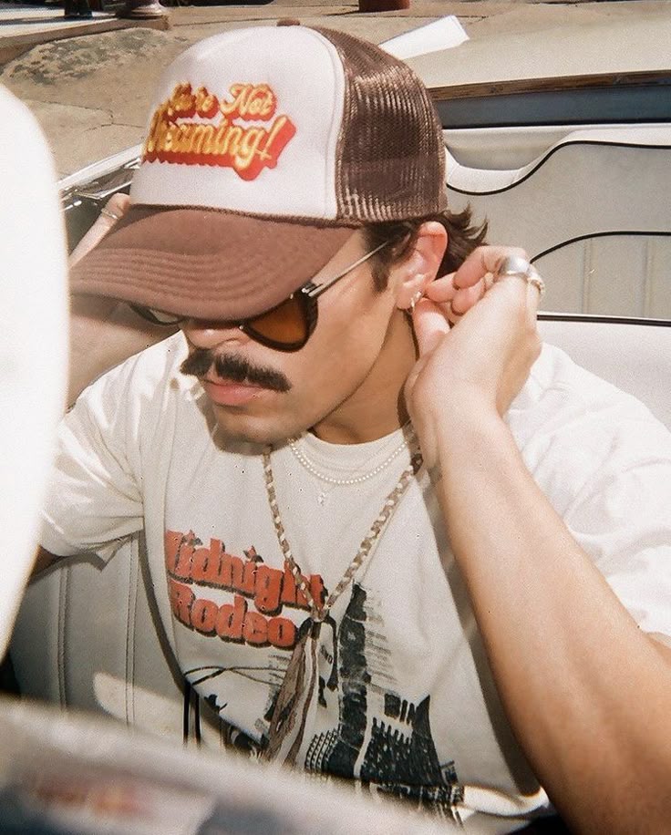 Man wearing a vintage cap and sunglasses in a car, adjusting his earring, captures retro style and fashion.