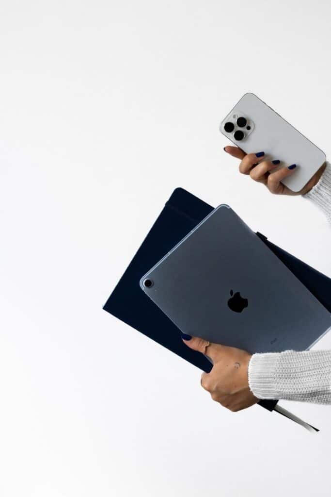 Person holding an iPhone, iPad, and notebook against a plain background.
