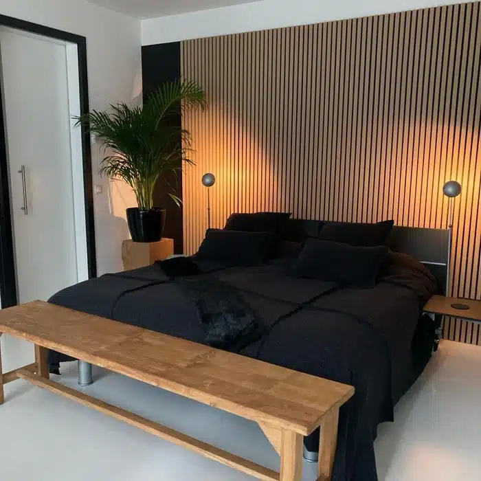 Modern bedroom with wooden slatted wall, cozy black bedding, bench, and indoor plant for a serene atmosphere.