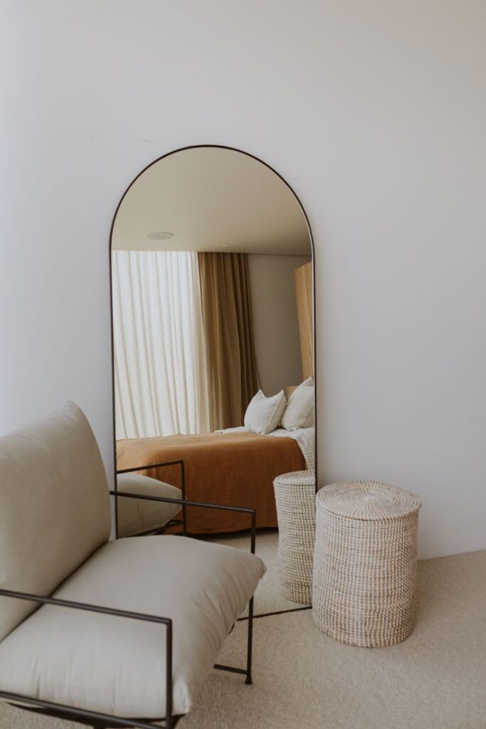 Modern bedroom with an arch mirror reflecting a cozy bed and neutral decor, beside a woven basket and plush chair.