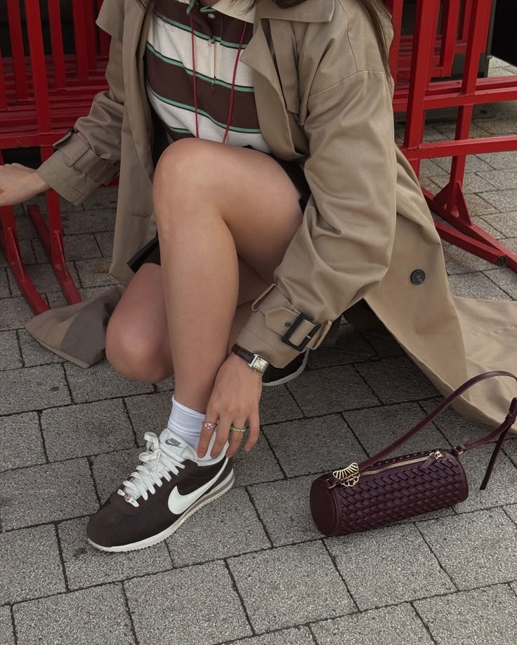 Fashionable outfit with trench coat, striped shirt, and Nike sneakers. Burgundy handbag in foreground. Urban setting.