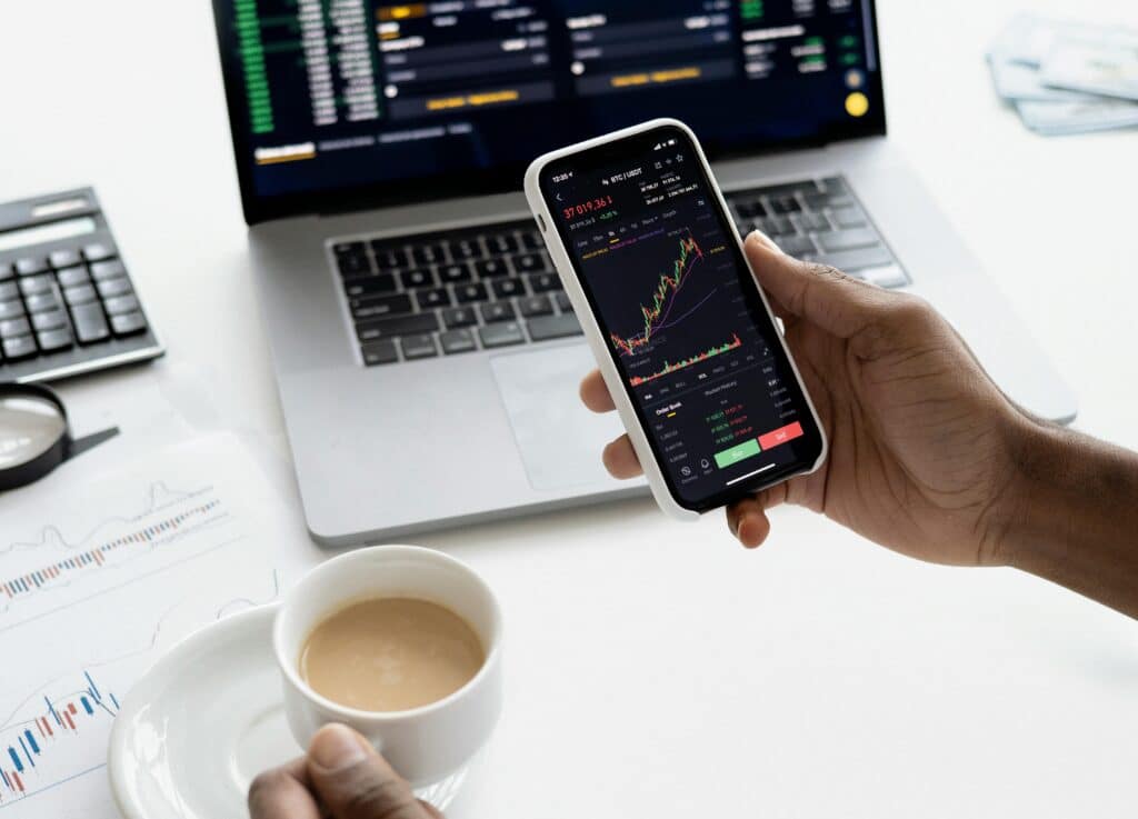 Person analyzing stock market charts on phone and laptop, holding coffee.
