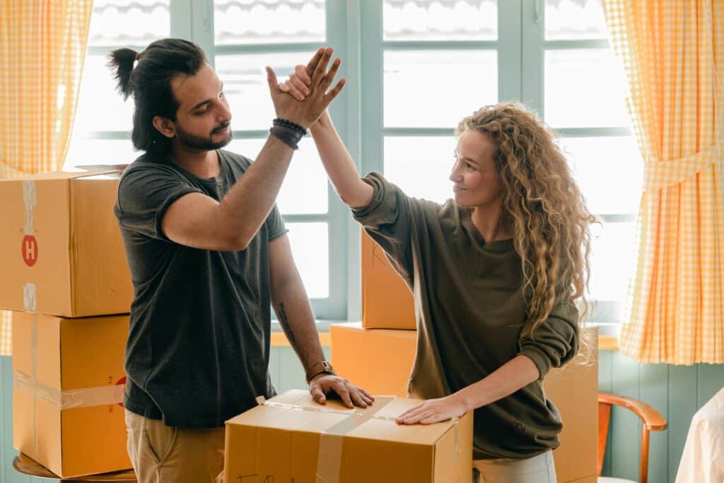 Couple high-fiving while unpacking boxes in a brightly lit room, celebrating a successful house move.