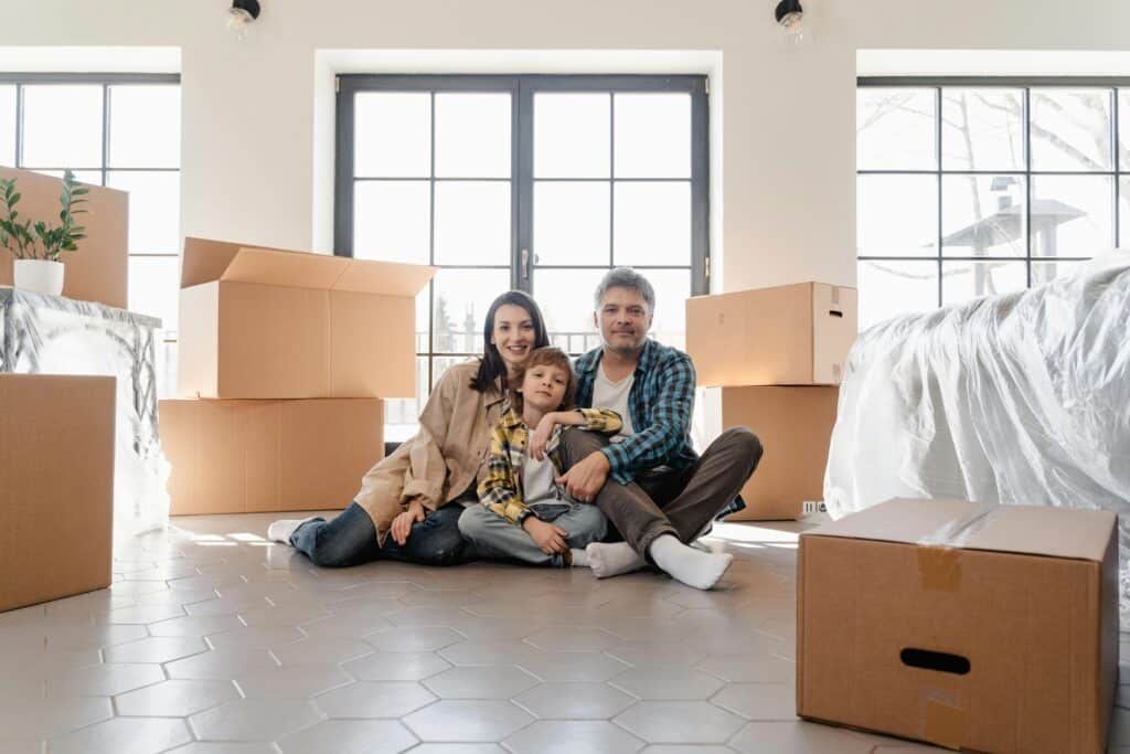 pexels-mart-production-7414919 Family sitting on floor surrounded by moving boxes, smiling in new home. | Sky Rye Design Family sitting on floor surrounded by moving boxes, smiling in new home.