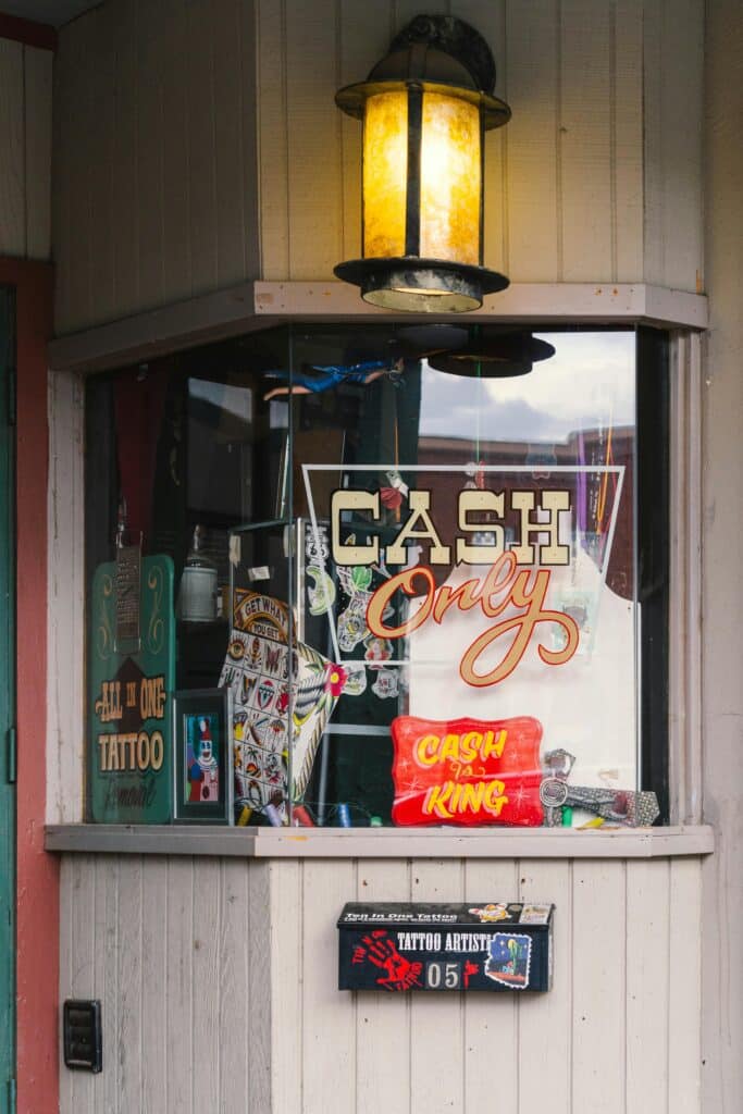 Tattoo shop window displaying Cash Only sign and art designs under a warm lantern light.