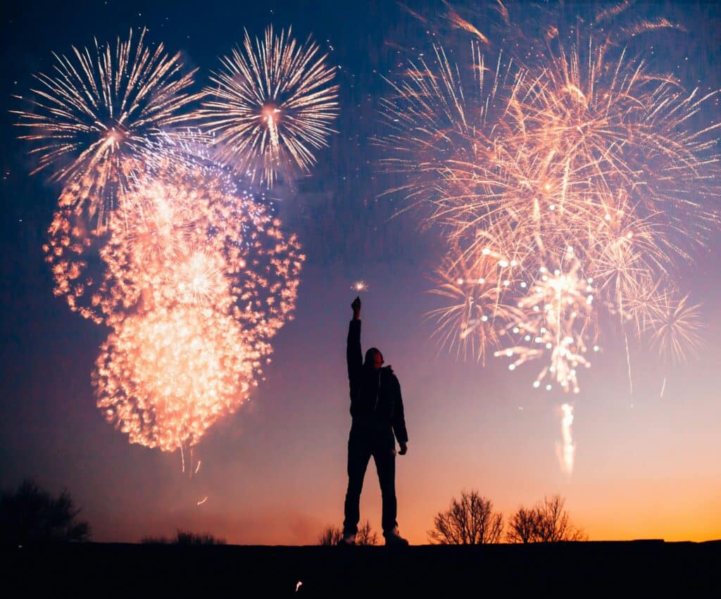 Silhouette of a person with sparkler watching vibrant fireworks against a twilight sky.