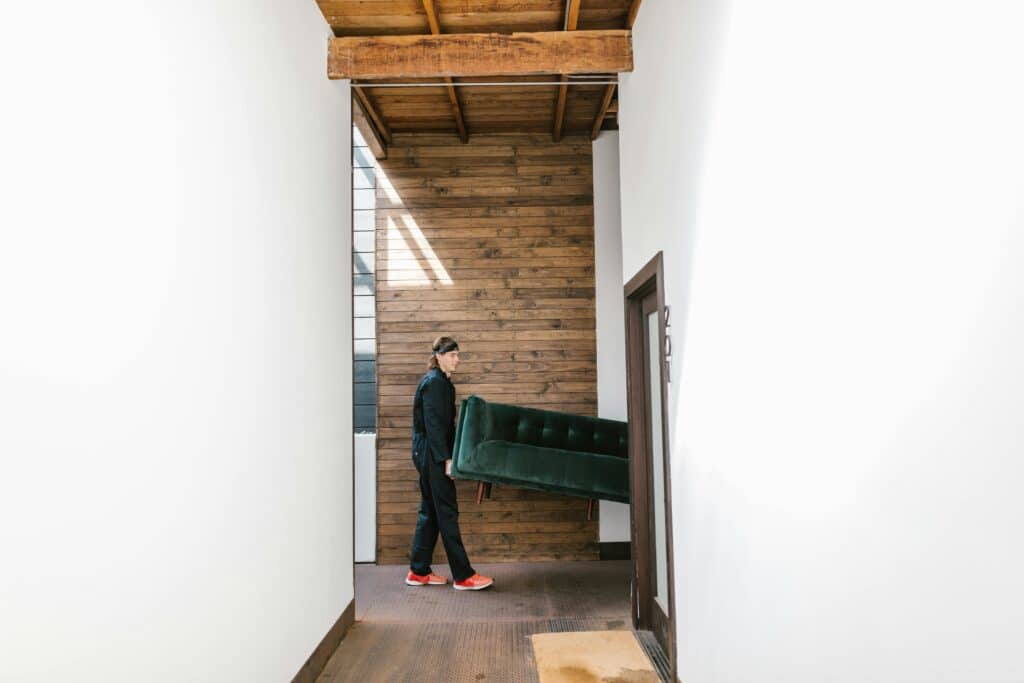 pexels-rdne-7464681 Person moving a green sofa through a hallway in a modern home with wooden accents and bright natural lighting. | Sky Rye Design Person moving a green sofa through a hallway in a modern home with wooden accents and bright natural lighting.