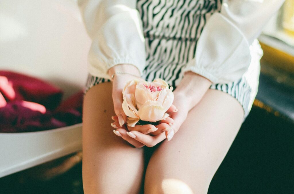 Woman holding a peach flower in hands, wearing a striped outfit, sitting by a window in soft natural light.