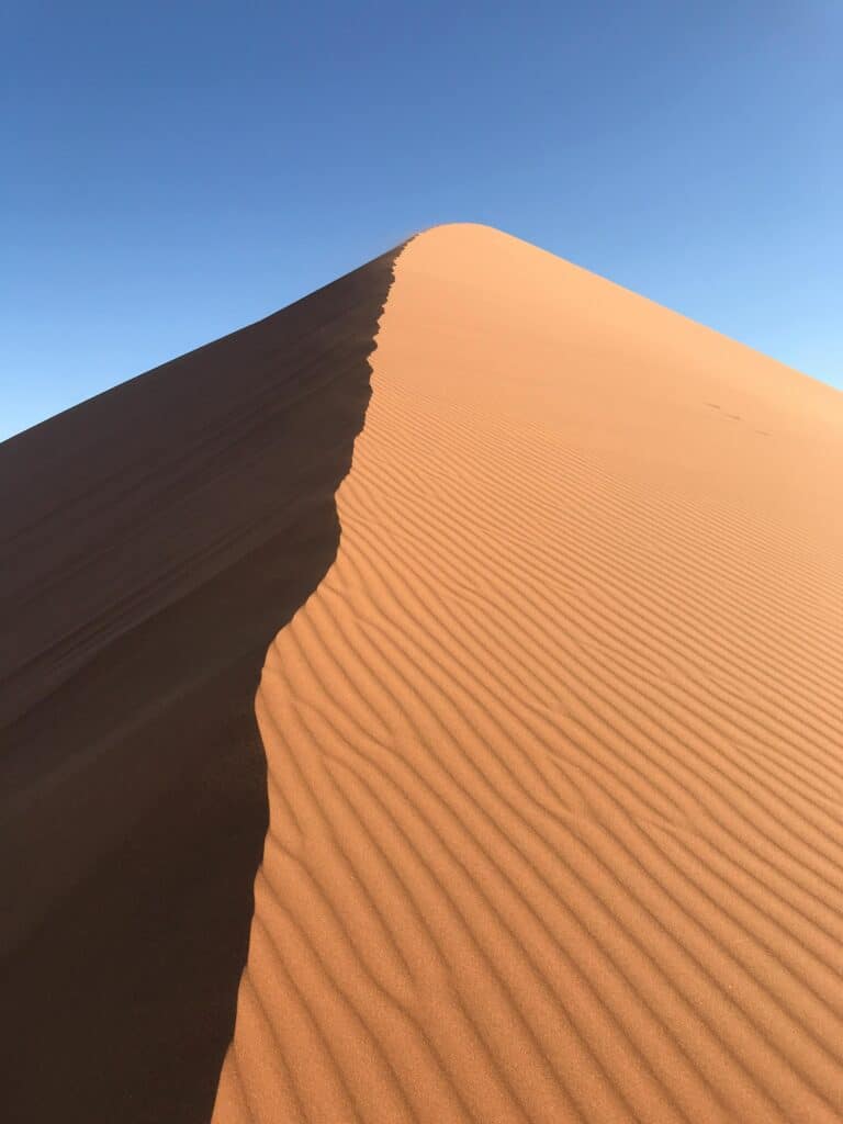 Sand dune with contrasting light and shadow under a clear blue sky, showcasing natural patterns and textures.