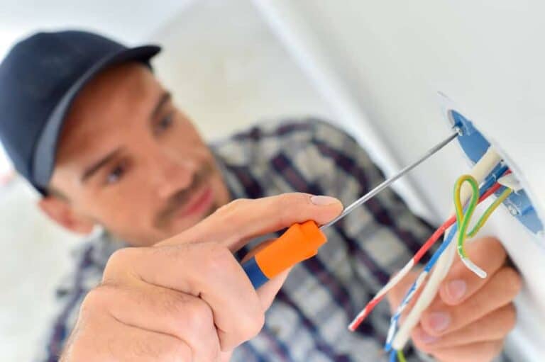 Electrician installing outlet with screwdriver and wires, wearing a plaid shirt and cap, focused on electrical work.