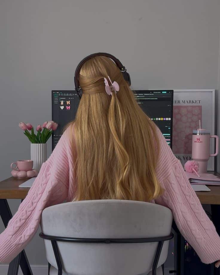 Young woman with headphones working at a computer desk, wearing a pink sweater, surrounded by pink decor.