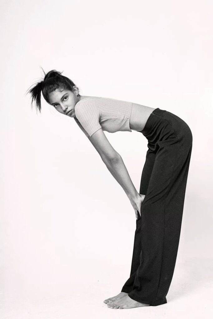 Woman in high-waisted pants and crop top bending forward, stylish fashion pose on white background.