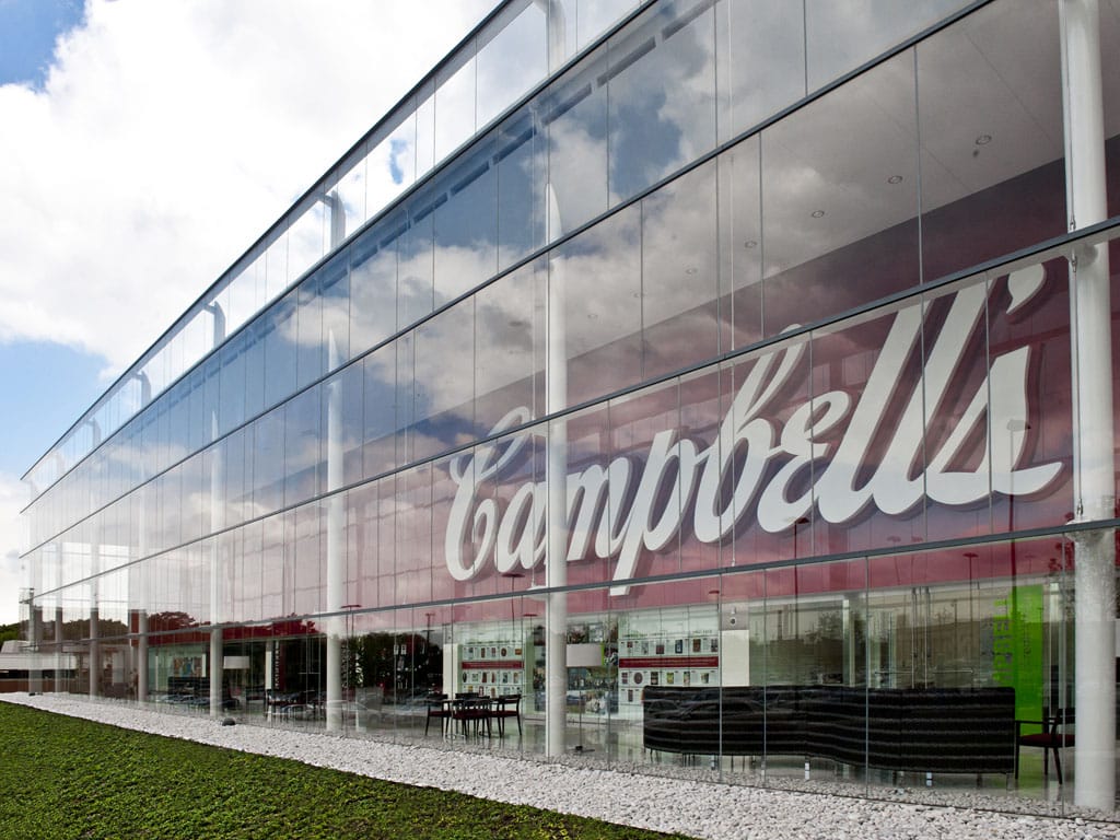 Modern building facade with reflective glass, featuring large Campbell's branding, under a partly cloudy sky.