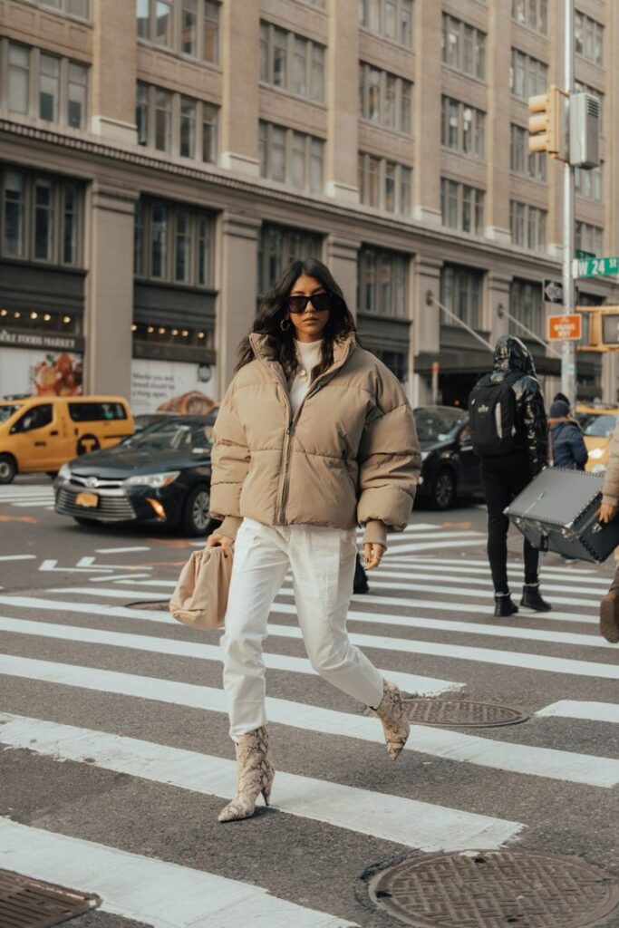 Person in stylish beige coat and white pants crossing a city street with taxis and buildings in the background.