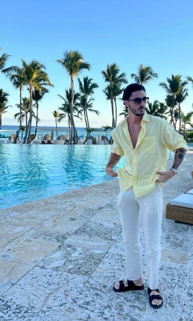Man in yellow shirt and white pants near tropical pool and palm trees under a clear blue sky.