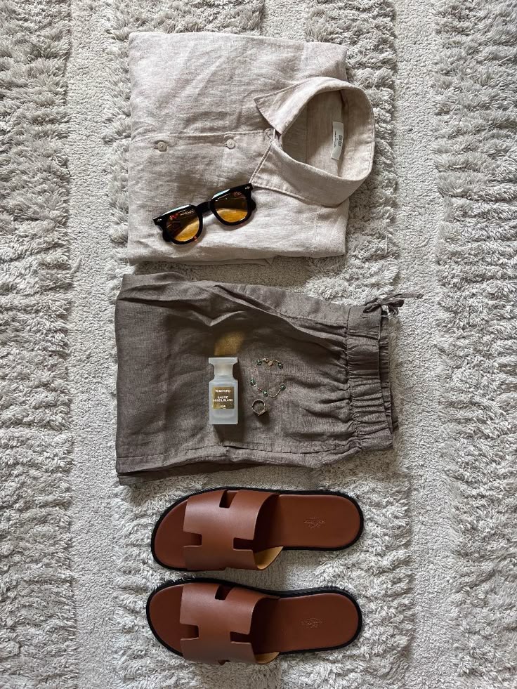 Flat lay of men's summer outfit: linen shirt, shorts, sunglasses, brown sandals, cologne, and accessories on a patterned rug.