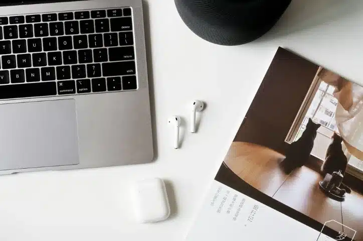 Laptop, wireless earbuds, and magazine on a white desk, featuring a cozy home office setup with tech and cats.