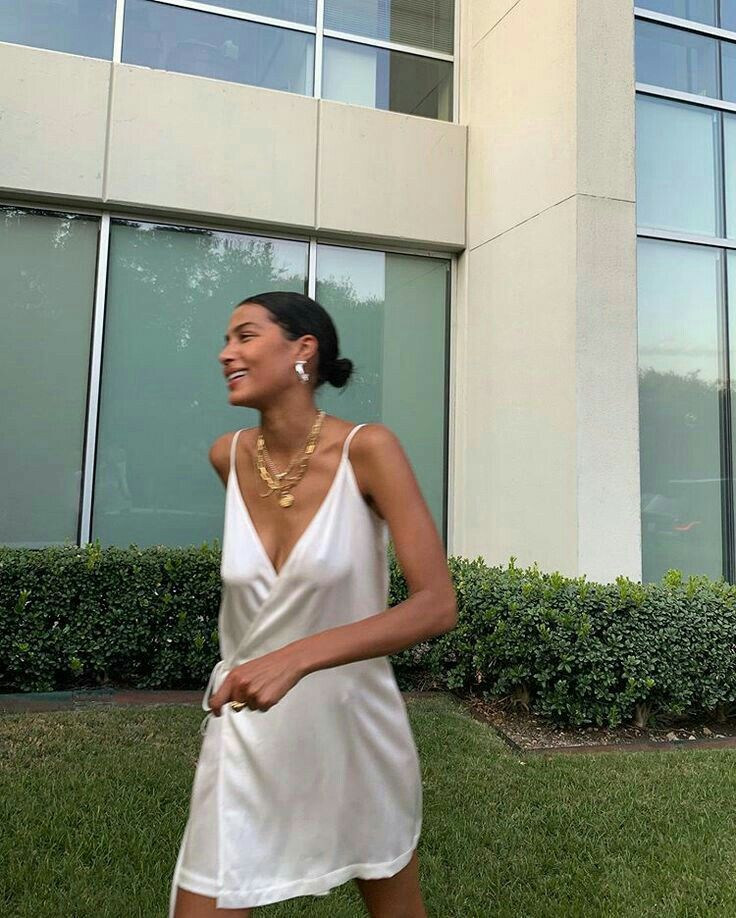 Woman in white dress with layered gold necklaces, smiling and walking outdoors beside modern building.