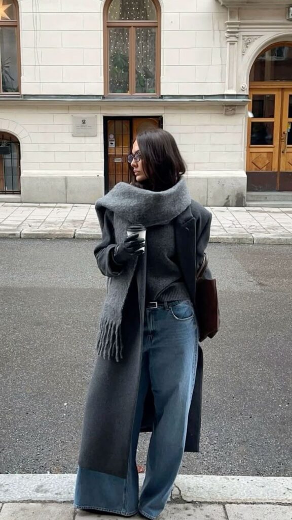 Stylish woman in gray coat and scarf holding coffee, standing on city street in winter fashion.