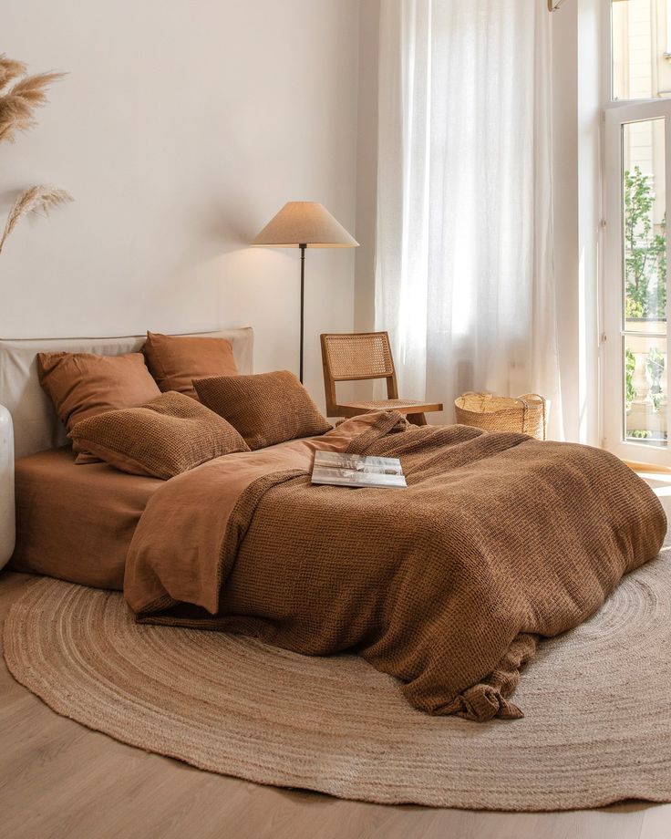 Cozy bedroom with brown bedding, wicker chair, and soft lighting. Natural decor and large window create a warm, serene atmosphere.