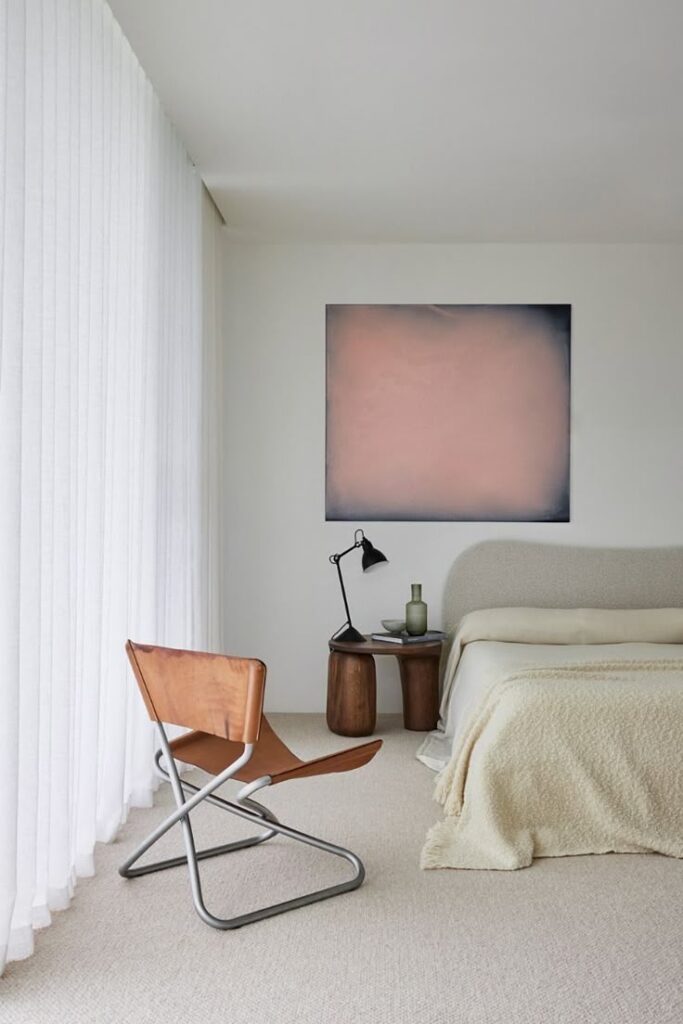 _ Modern minimalist bedroom with neutral tones, featuring a leather chair, wooden side table, and abstract wall art. | Sky Rye Design Modern minimalist bedroom with neutral tones, featuring a leather chair, wooden side table, and abstract wall art.