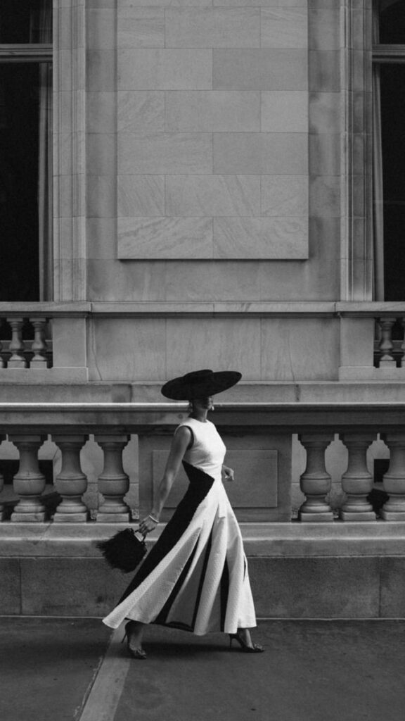_ Elegant woman in a wide-brim hat and stylish dress walking past a grand building in black and white. | Sky Rye Design Elegant woman in a wide-brim hat and stylish dress walking past a grand building in black and white.
