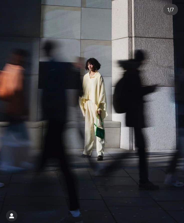 _ Person in yellow outfit stands in urban setting amid blurred pedestrians, holding green bag, with sunlight casting shadows. | Sky Rye Design Person in yellow outfit stands in urban setting amid blurred pedestrians, holding green bag, with sunlight casting shadows.