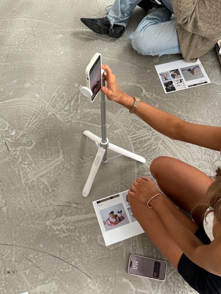 Person taking photo with smartphone on tripod, sitting on floor with brochure and phone.