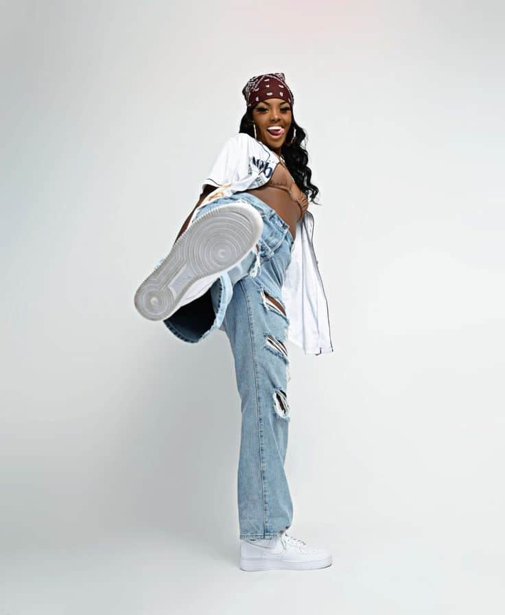 Fashionable woman kicks high, wearing ripped jeans, a bandana, and white sneakers against a white background.