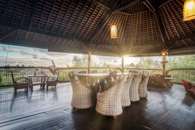 Open-air pavilion with wicker chairs and scenic view of greenery and sunset. Ideal for dining and relaxation.
