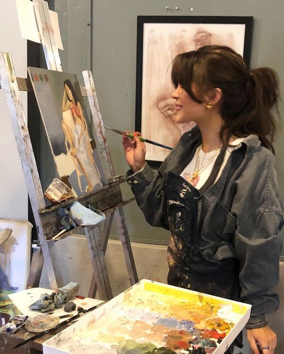 Artist painting on canvas in studio, wearing denim shirt and apron, with colorful palette and art supplies nearby.