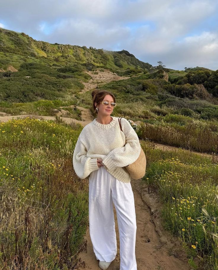 Woman in cozy sweater and white pants walking through a scenic green meadow with a straw bag.