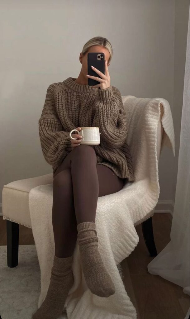 Cozy woman in brown sweater and leggings, sitting on chair with mug, taking a mirror selfie.