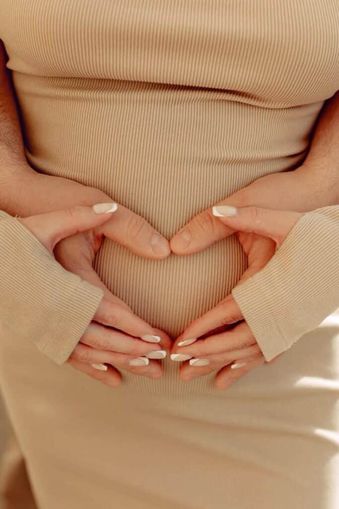 Pregnant woman in beige dress with hands forming heart on belly, symbolizing love and anticipation of new life.