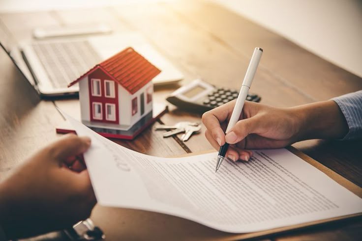 Person signing real estate contract with miniature house, keys, and calculator on table.