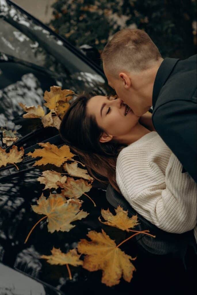 Couple embraces on leaf-covered car hood in autumn, sharing a tender moment.