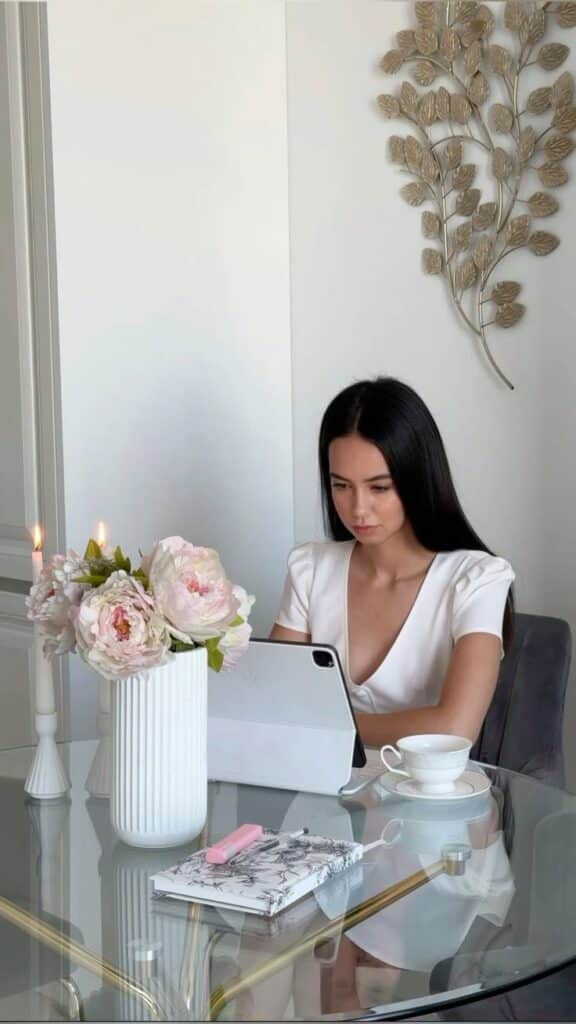 Woman working on a tablet at a glass table with flowers, a notebook, and a teacup in a stylish home office.