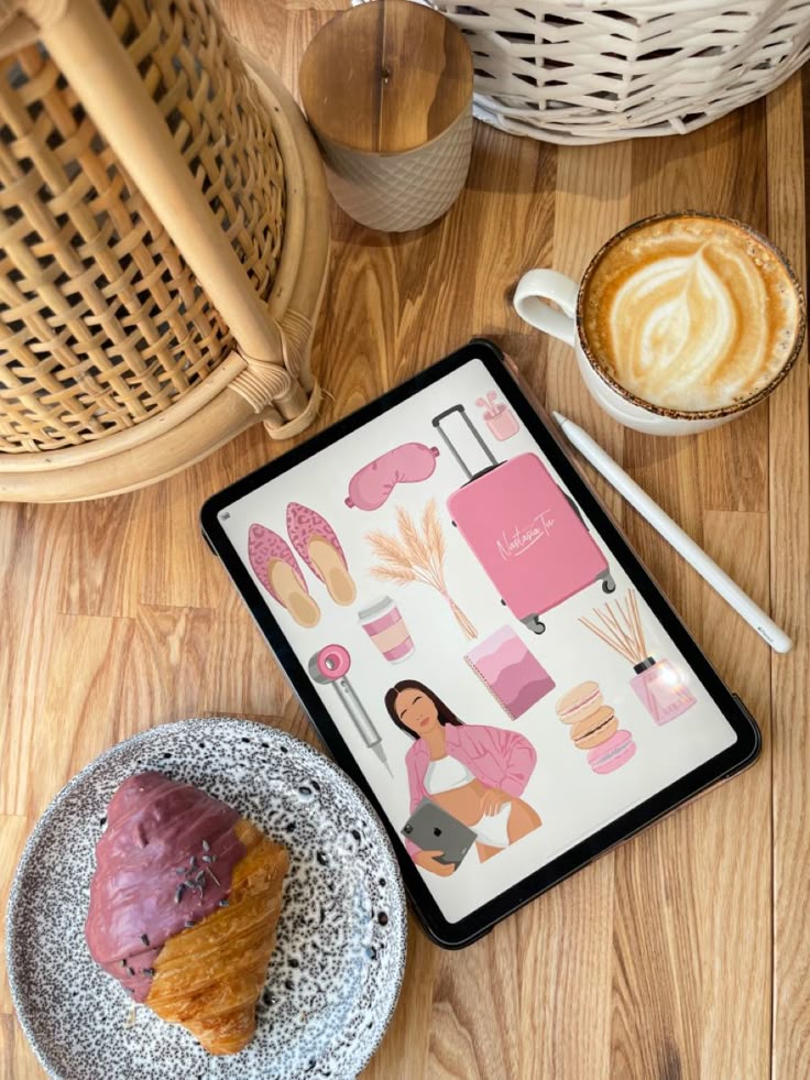 Tablet with pink-themed illustrations, next to a latte and a croissant on a wooden table. Cozy ambience.