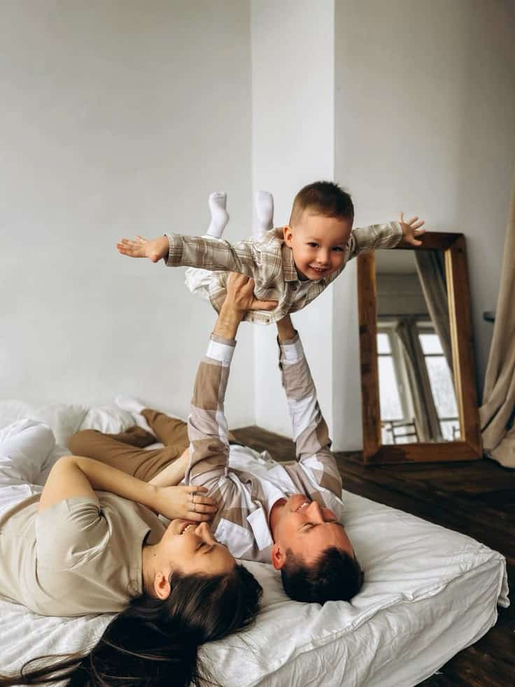 A joyful family enjoys playtime on a bed, with parents lifting their smiling child in the air.