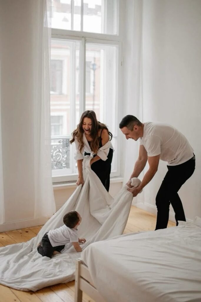 Family enjoying playful time making the bed, smiling in a bright bedroom with white sheets.