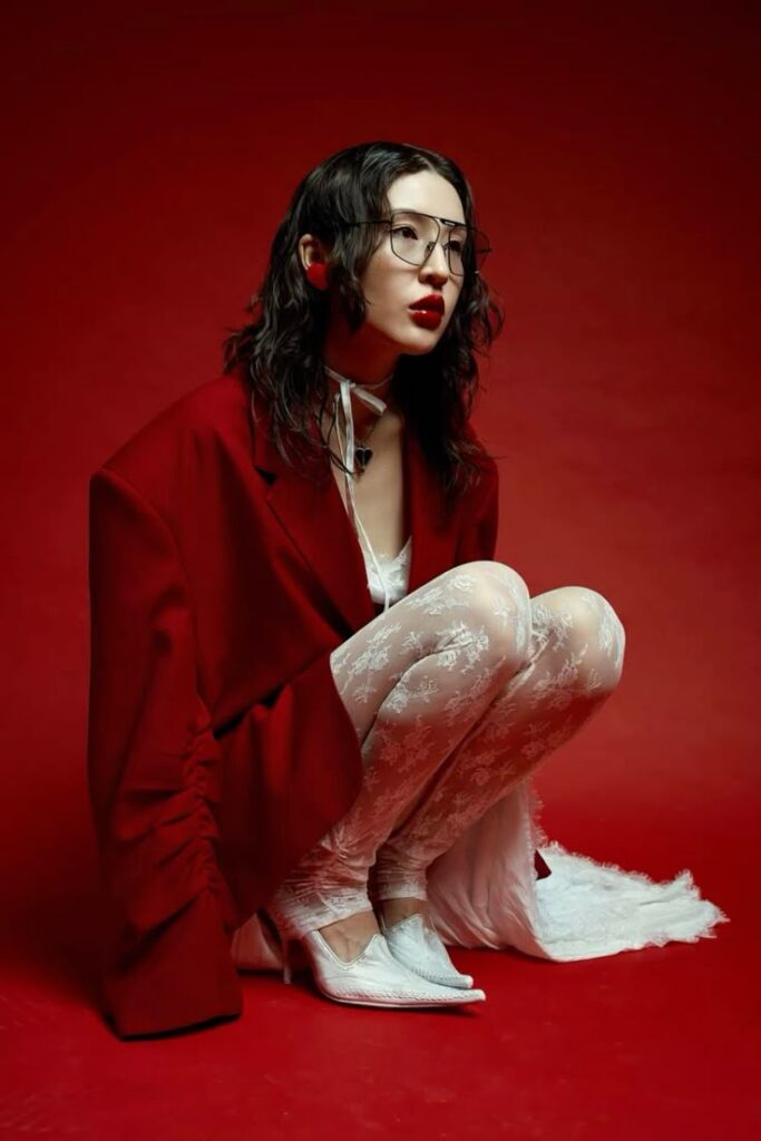 Fashionable woman in red blazer and lace outfit against red background, wearing glasses and white heels.