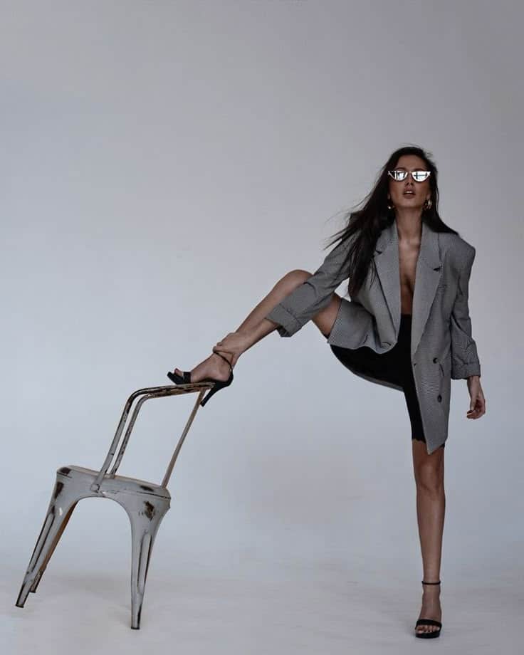 Fashion model posing creatively with chair, wearing stylish blazer and sunglasses, minimalistic background.