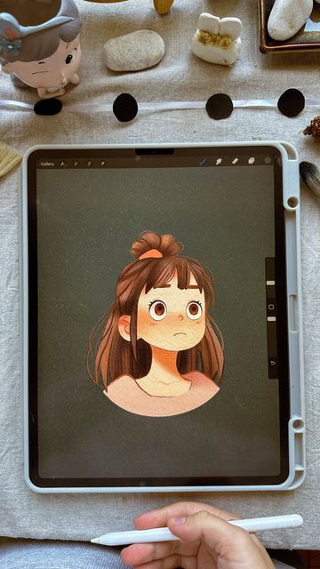 Digital artwork of a girl with a top bun displayed on a tablet, surrounded by art supplies and figurines.