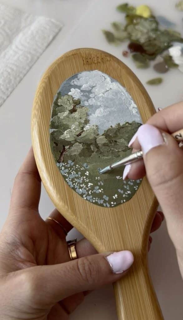 Hand painting a landscape on a wooden hairbrush using delicate brush strokes; artistic DIY project.