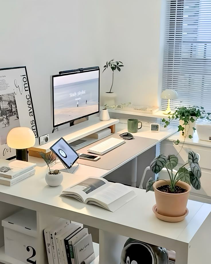 Minimalist home office setup with books, plants, and a computer on a white desk beside a window with blinds.