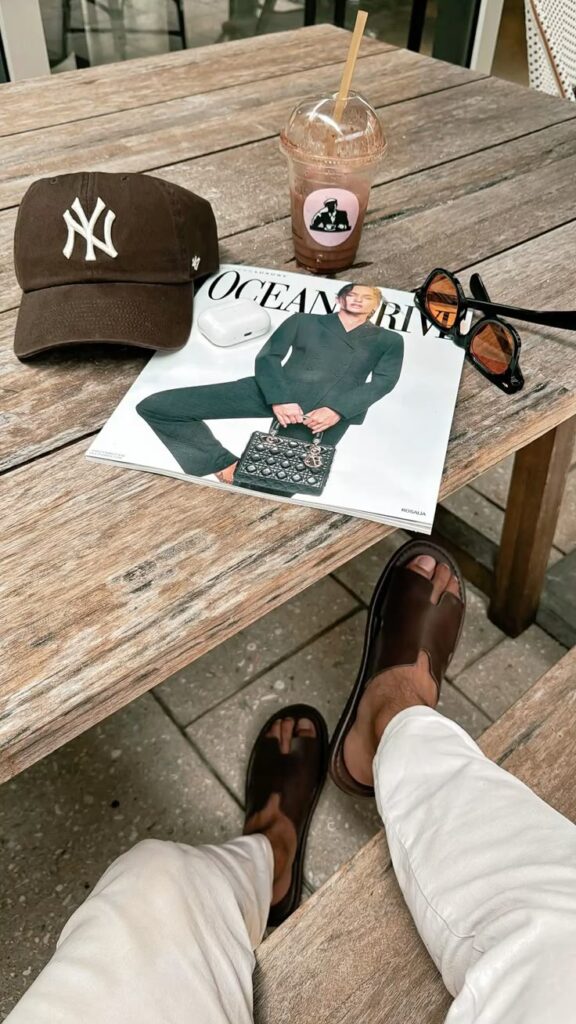 Casual outdoor table with magazine, cap, sunglasses, iced coffee, earphones, and sandals. Relaxed lifestyle scene.